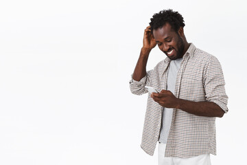 Happy guy receive message from his date in app, smiling and blushing pleased, touching head. Handsome african-american bearded man using smartphone, look mobile display and grinning, white background