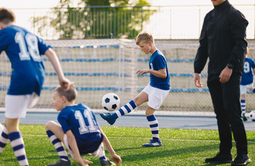 Wall Mural - Boy juggling soccer ball. Happy children playing sports on grass turf field. Group of kids on training with coach. Children in blue soccer jersey on practice session