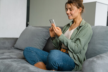 Wall Mural - Happy nice woman using mobile phone while sitting on sofa