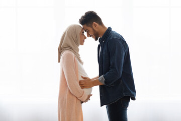 Wall Mural - Loving pregnant muslim couple bonding together at home, standing near window