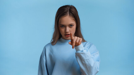 Wall Mural - teenage girl in sweatshirt pointing with finger while looking at camera isolated on blue