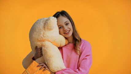 Wall Mural - happy teenage girl in pink sweatshirt holding teddy bear isolated on yellow