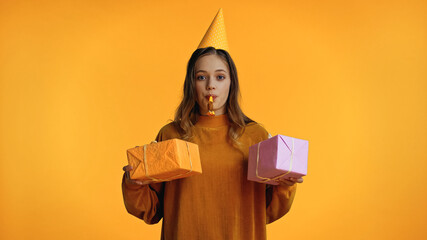 Wall Mural - teenage girl in party cap looking at camera while blowing party horn and holding presents isolated on yellow