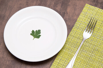 Single one parsley green leaf on white empty plate and fork on table. Diet, fasting, vegan, vegetarian, healthy food concept