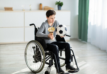 Teen handicapped soccer player with ball and trophy sitting in wheelchair, feeling depressed over his injury at home