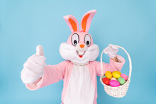 Happy Easter. Easter Bunny Or Rabbit Or Hare Holds Egg With Basket Of Colored Eggs, Having Fun, Celebrates Easter Rabbit Isolated On Blue Background