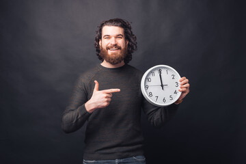 Wall Mural - Cheerful handsome bearded man in casual pointing at clock over black background.