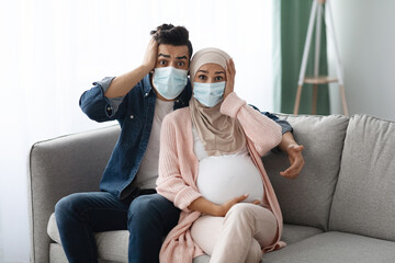 Wall Mural - Pregnancy During Pandemic. Shocked Pregnant Muslim Couple In Masks Sitting On Couch