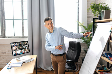 Wall Mural - Middle aged successful male top manager or business tutor standing near flipchart in modern office, conducts an online lesson or presentation. Satisfied businessman stylishly dressed on video meeting