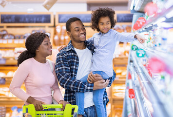 Wall Mural - Happy black parents and their pretty daughter with shopping cart having fun buying groceries at hypermarket