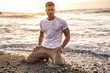 Gorgeous male model in soaked wet t-shirt posing on the beach, with sunset ocean on the background.