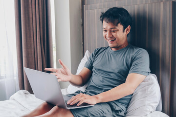 Happy Asian man is working with his laptop on his bed. Concept of freelancer successful lifestyle.