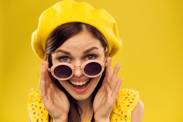 Happy woman in a yellow hat and fashionable glasses holds her hands near her face on a yellow background cropped view of emotions fun