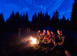 © anatoliy_gleb - Company of young people is sitting around the fire against the backdrop of the silhouetted of large spruce trees and dark blue night sky. Evening camping in the forest. Vacation and tourism concept