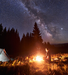 Wall Mural - side view of female tourist having a rest by campfire on background of tent and spruce forest under 