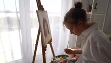 Young Artist Paints A Picture With A Brush On Canvas Sitting Near The Window In The Studio. Teenage Girl Painting A Self-portrait