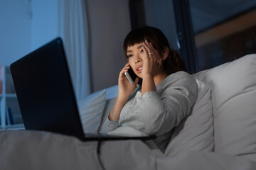 technology, internet, communication and people concept - stressed young asian woman with laptop computer calling on smartphone in bed at home at night