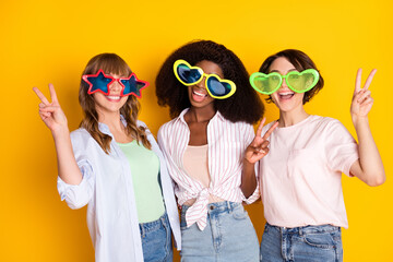 Sticker - Portrait of attractive trendy cheerful girls embracing showing v-sign wearing large specs isolated over bright yellow color background