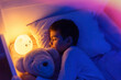 © Elena - Little preschool kid boy sleeping in bed with colorful night led lamp. School child dreaming and holding plush toy. Kid angry of darkness.