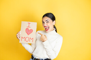 Wall Mural - Beautiful woman celebrating mothers day holding poster love mom message amazed and pointing with hand and finger