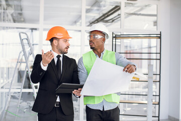 African and caucasian men in protective helmets using digital tablet and blueprints while controlling working process on construction. Cooperation concept.