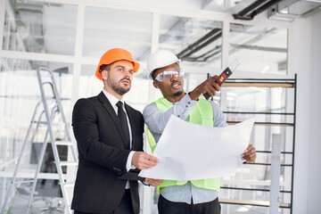 African and caucasian men in protective helmets using digital tablet and blueprints while controlling working process on construction. Cooperation concept.