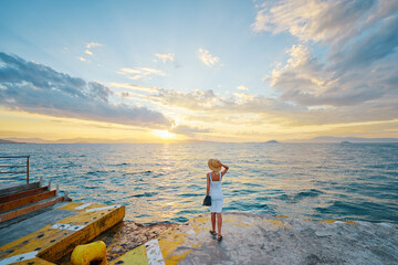 Wall Mural - Holiday in Greece. Woman in hat enjoying beautiful sunset sea view from wharf.