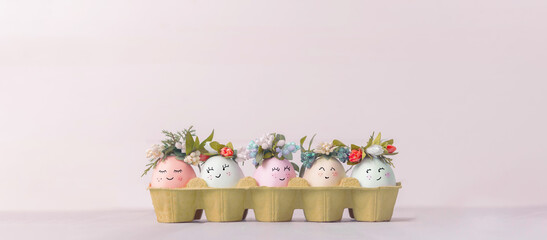 Wall Mural - Easter concept. Easter eggs painted in pastel colors with cute faces in an egg tray with wreaths put on them. Light background, place for text.