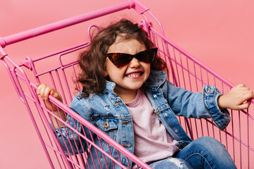 Blissful preschool child sitting in shopping cart. Laughing kid in jeans having fun on pink background.