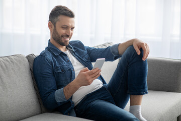 Wall Mural - Happy man sitting on couch at home, using mobile phone