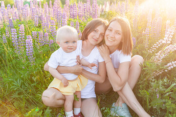 Young mother embracing her kids outdoor. Woman baby child and teenage girl sitting on summer field with blooming wild flowers green background. Happy family mom and daughters playing on meadow.