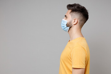 Wall Mural - Side profile view of young caucasian man 20s wearing yellow t-shirt in sterile face mask to safe coronavirus virus covid-19 pandemic quarantine look aside isolated on grey background studio portrait.