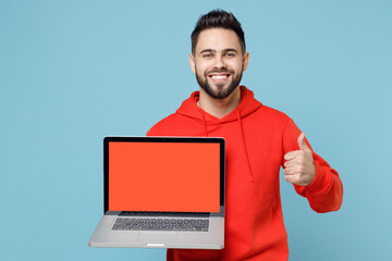 Wall Mural - Young caucasian smiling web designer bearded man 20s in casual red orange hoodie hold laptop pc computer, blank screen workspace area show thumb up gesture isolated on blue background studio portrait.
