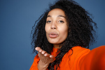 Wall Mural - Close up of pretty young african american woman in casual basic bright orange sweatshirt doing selfie shot on mobile phone blowing sending air kiss isolated on blue color background studio portrait.