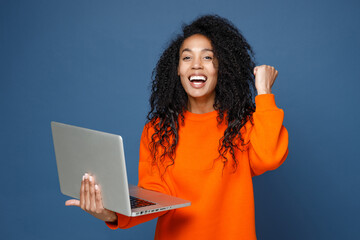 Wall Mural - Happy joyful young african american woman wearing casual basic bright orange sweatshirt standing working on laptop pc computer doing winner gesture isolated on blue color background studio portrait.