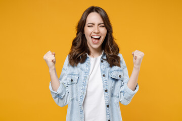 Wall Mural - Young excited overjoyed happy fun brunette expressive woman in stylish casual denim shirt white t-shirt do winner gesture clench fist celebrate isolated on yellow color background studio portrait.