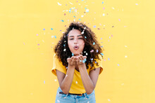 Young Woman Blowing Multi Colored Confetti Against Yellow Wall