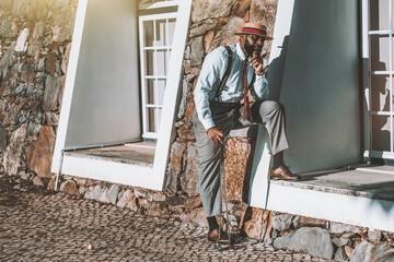 A portrait of a fashionable mature black guy with a beard and in a hat and an elegant costume waiting for his friends before a golf play while standing with a cigar in hand next to the building facade