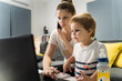 © Miljan Živković - Front view portrait of small caucasian boy looking to the laptop while sitting with his mother working on the computer by table on the sofa couch at home making a phone call work from home concept