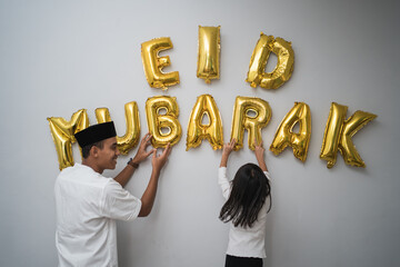 portrait father and daughter muslim decorating eid mubarak letter made of baloon decoration against the wall at home