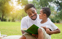 Cheerful African American Boy And Dad Having A Picnic In The Park, Happy Son And Father Reading A Book, Happiness Family Concepts