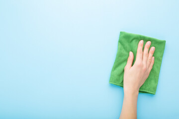 Young adult woman hand holding green rag and wiping table, wall or floor surface in kitchen, bathroom or other room. Closeup. Empty place for text or logo. Light pastel blue background. Top down view.