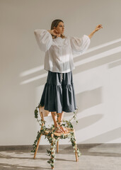 woman standing barefoot on a wooden chair with legs tied with green twigs with leaves.