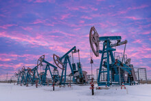 Under The Purple-blue Sky With Clouds Seven Oil Pumpjack Winter Working. Oil Rig Energy Industrial Machine For Petroleum In The Sunset Background For Design.