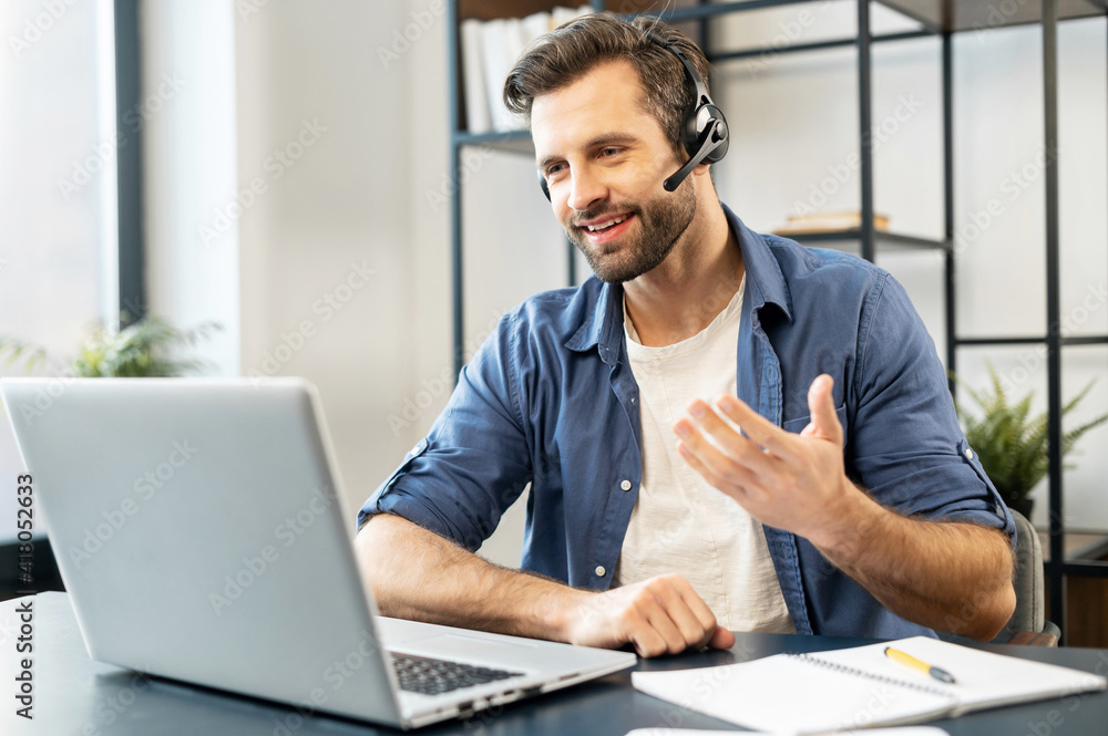 Confident man with handsome stubble, wearing headset, talking in front of computer, wearing casual clothes, talking at computer, discussing work process, gesticulates, chatting with partners, notice - obrazy, fototapety, plakaty 