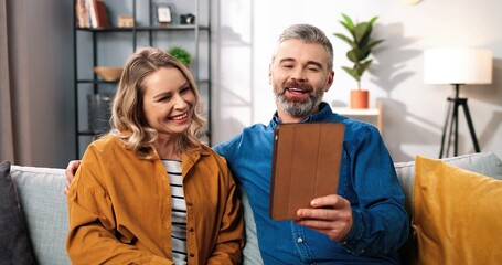 Wall Mural - Portrait of happy joyful Caucasian couple man and woman videochatting online on tablet in good mood, middle-aged wife and husband having internet conversation on device at home, leisure concept