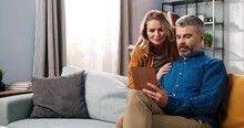 Portrait Of Handsome Caucasian Middle-aged Bearded Grey-haired Man Sitting On Sofa In Room Surfing Internet On Tablet Browsing And Choosing Something With His Beautiful Happy Wife, Family Concept