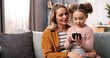 © VAKSMANV - Close up portrait of happy Caucasian female parent mom sitting on couch in living room at home with little cute daughter tapping on cellphone surfing internet together, parenting concept