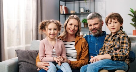 Wall Mural - Portrait of cheerful positive Caucasian family parents with children gathered together at home sitting on sofa in room looking at camera and smiling in good mood, mom, dad with small daughter and son