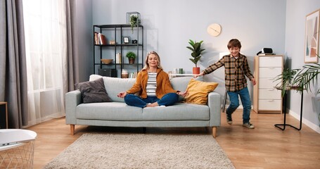 Wall Mural - Beautiful Caucasian mom parent sitting on couch meditating with calm face and closed eyes in living room while her little child boy running around playing and annoying her, meditation concept